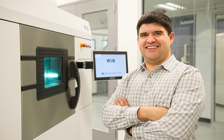 Victor Lopez in standing and smiling in front of an EOS 3D printing