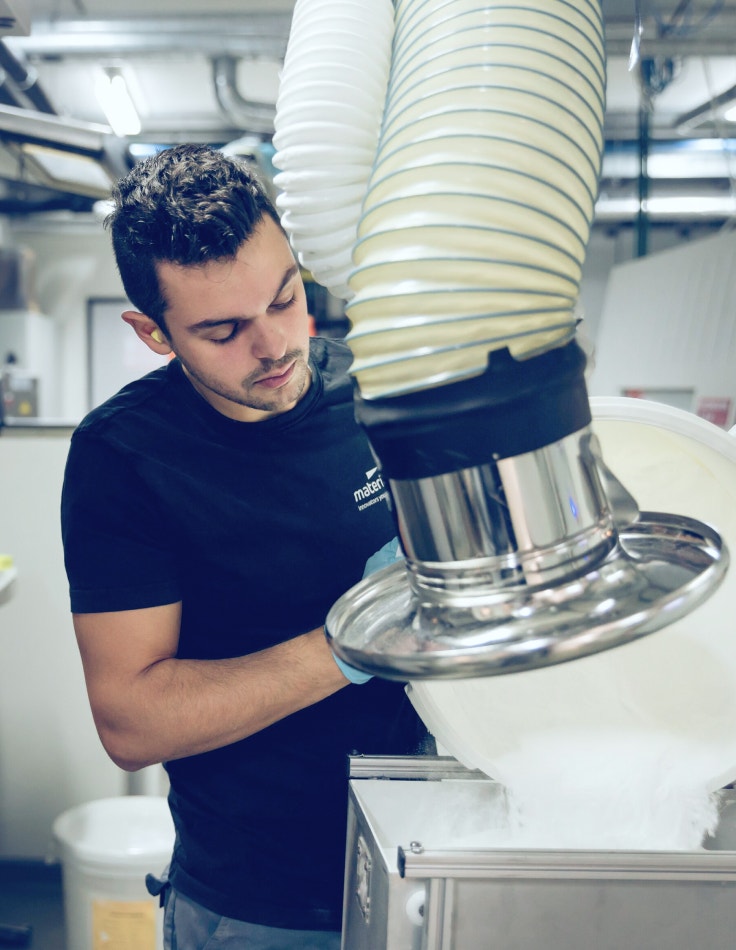 A 3D printing production worker is emptying a bucket of white PA 12 powder and small 3D-printed parts into a metal container. A large suction nozzle above the container draws up the loose powder, leaving the laser sintered parts behind for further cleaning.