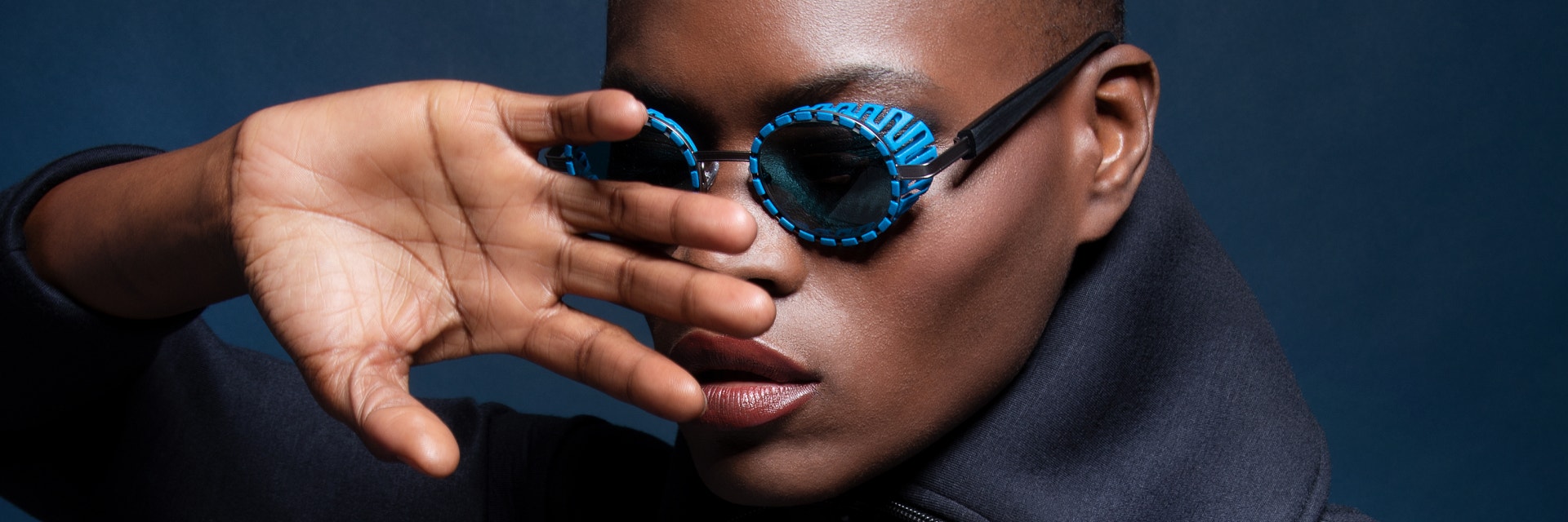 Black female model posing with her hand in front of her face while wearing blue sunglasses from the JFRey Nautinew collection