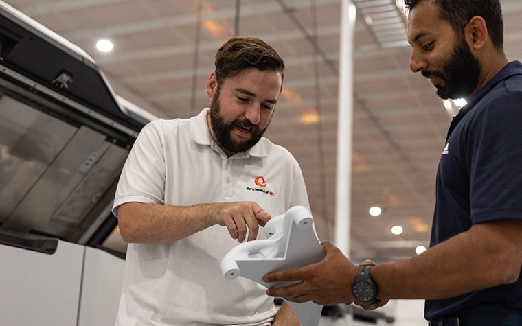 An Endeavor 3D team member holding a white 3D-printed part whild another team member points to a component of the part