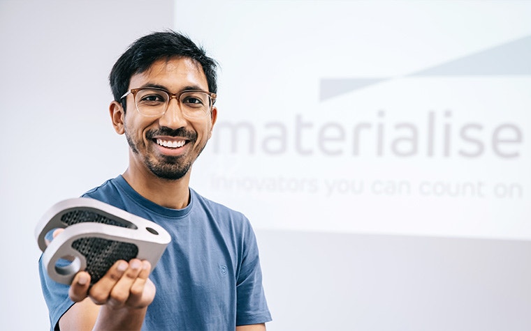 Man standing and smiling in front of a screen projecting the Materialise logo, holding a metal 3D-printed part