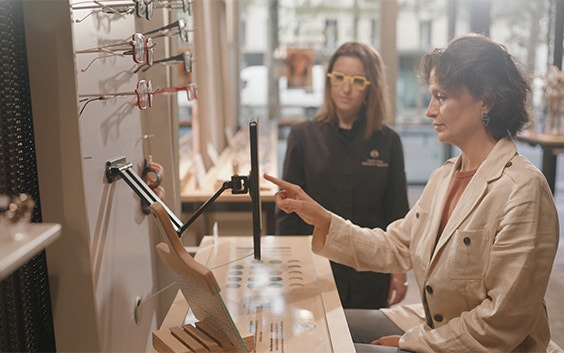A woman uses the Materialise Eyewear Fitting Suite on an iPad in an Acuitis store, watched by her optician.