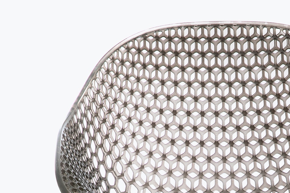 Close-up view of a geometrical, 3D-printed chair seat from a 3D-printed prototype of a customized, futuristic-looking wheelchair, using multiple 3D printing materials. The seat is lattice-structured and made of a translucent gray resin.