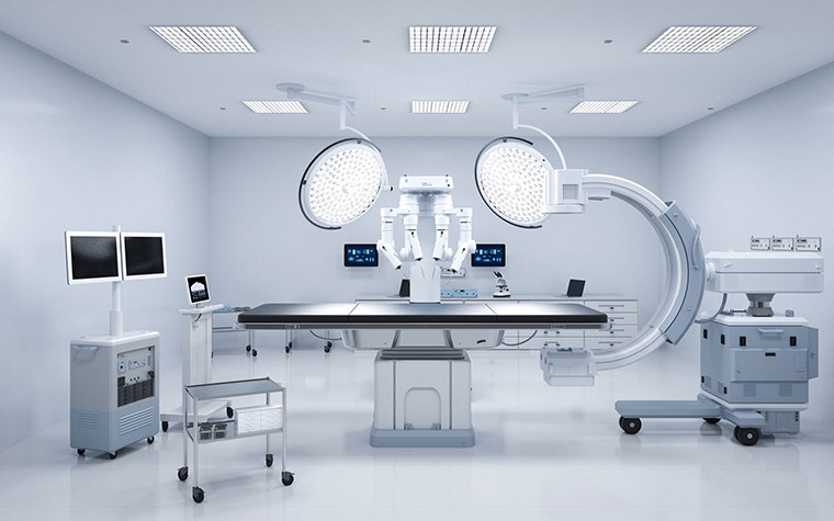 A modern operating room with advanced medical equipment, including large overhead surgical lights, a robotic surgical system, dual monitors, and various mobile carts, all arranged around a central operating table in a sterile, brightly lit environment.