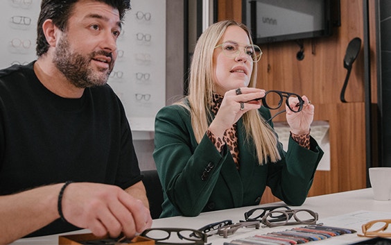 Two people holding up 3D-printed eyewear frames at a table with many 3D-printed frames around