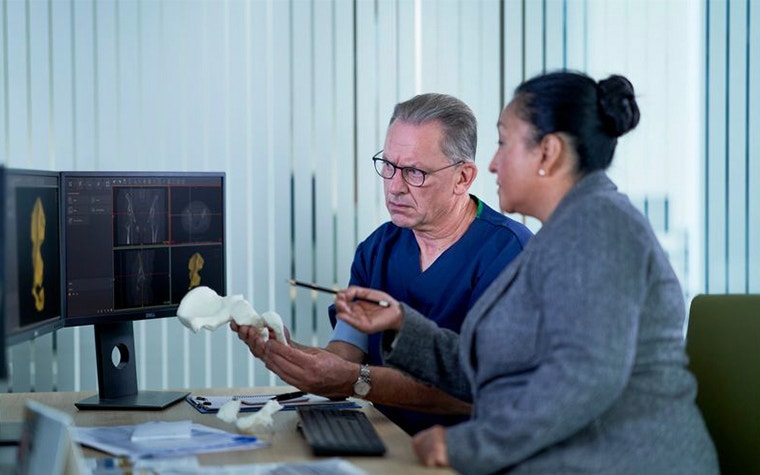 A radiologist and surgeon look at computer screen with 3D anatomical image of hip bone on it 