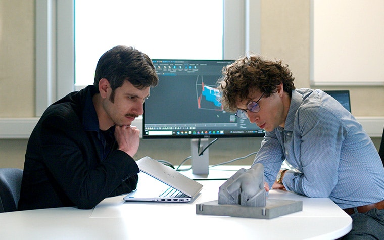 Two men sitting at a desk looking at metal 3D-printed part with thin support structures.