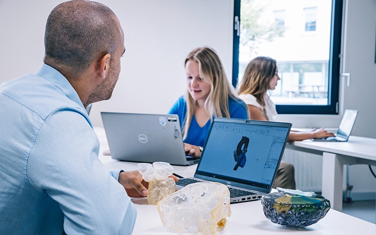 A trainee sits at a laptop, looking at an anatomical model in Mimics and 3D-printed anatomical models. Two trainees sit in the background on their laptops