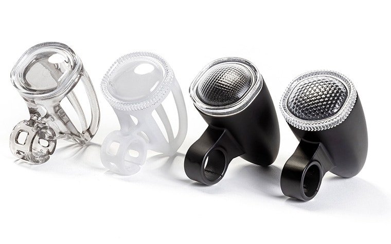 Four 3D-printed bicycle light housings made with VersaClear displayed in a row, showing different shades, from transparent and white prototypes to finished black units with textured reflective lenses.