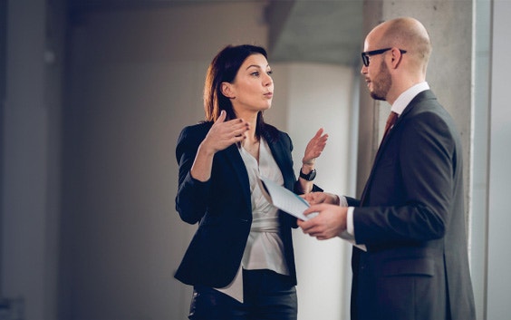 Two business people are talking in a corporate environment. The woman on the left is gesturing with her hands and the man on the right is holding a paper folder.