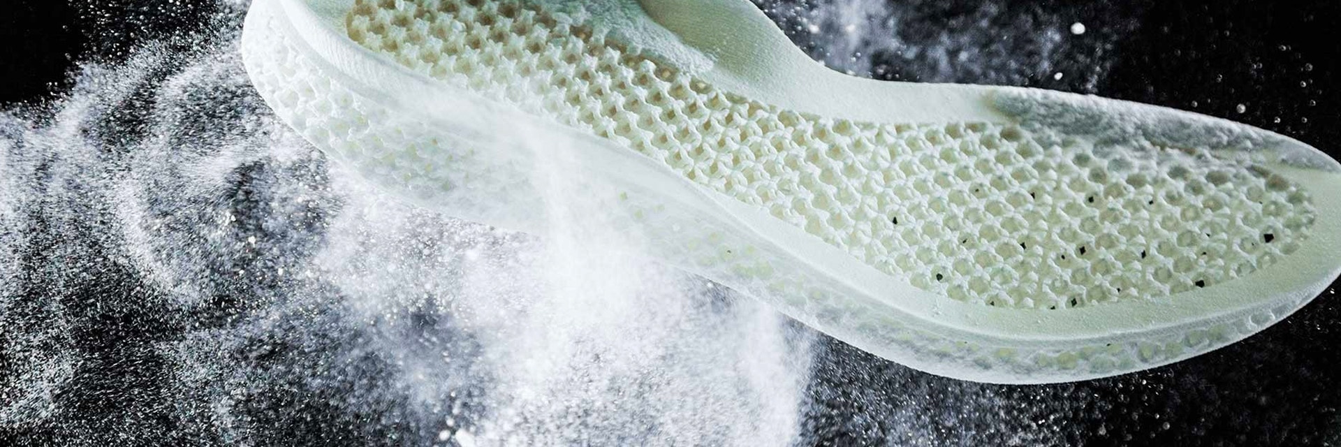 A 3D printed Adidas midsole is shown tossed up in mid-air surrounded by a cloud of white PA 12 powder. The midsole has a lattice structured base typical of laser sintering.