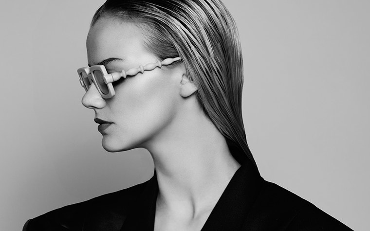 Immagine in scala di grigi di una modella con capelli sciolti che guarda di lato e indossa occhiali BAARS x Gogosha di colore nudo.