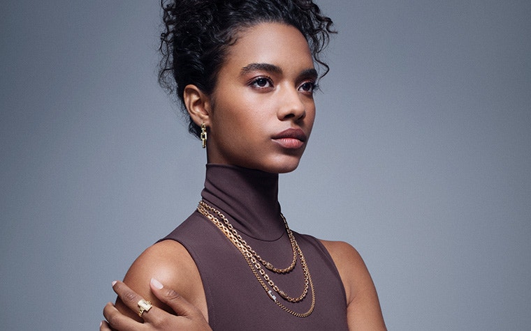 ノースリーブのシャツを着て右を見つめる女性。手は肩に触れている。首には細い金の鎖が3本、人差し指には金の指輪。