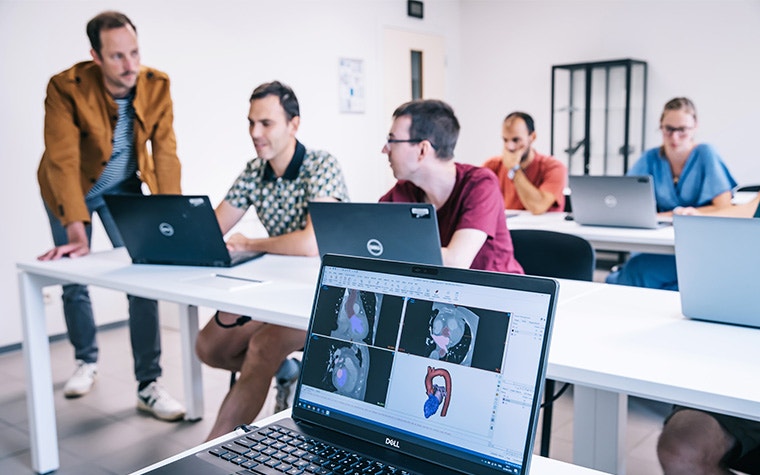 In the foreground is a laptop, showing Mimics software with images and models of patient anatomy. In the background is a Materialise trainer speaking with trainees sitting at a table.