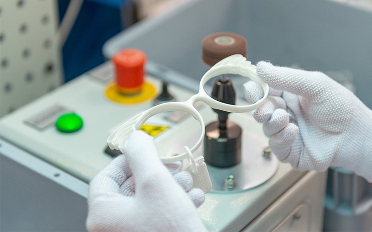 Two gloved hands holding white, 3D-printed eyewear frames above a control panel