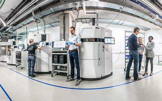 Panoramic view of a production facility with people speaking and checking on 3D printers