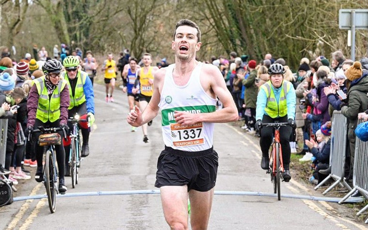 Distance runner Dan Studley crosses the finish line in the 2023 Trafford 10k, followed by three people on bicycles wearing high-vis jackets. A crowd of spectators stand on either side.