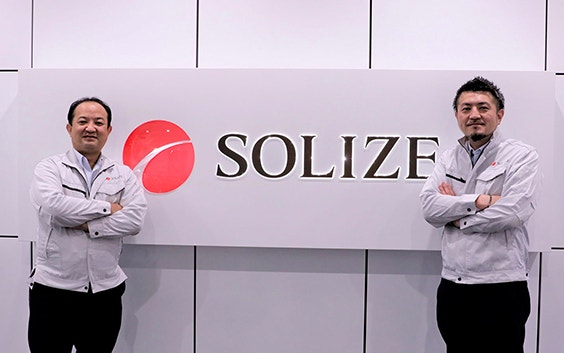 SOLIZEの看板の横に立つSOLIZEチームメンバー2名。