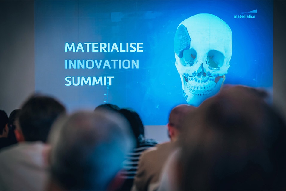 A screen showing a slide with the words "Materialise Innovation Summit" and a skull model with personalized implants attached in front of a crowd of event attendees
