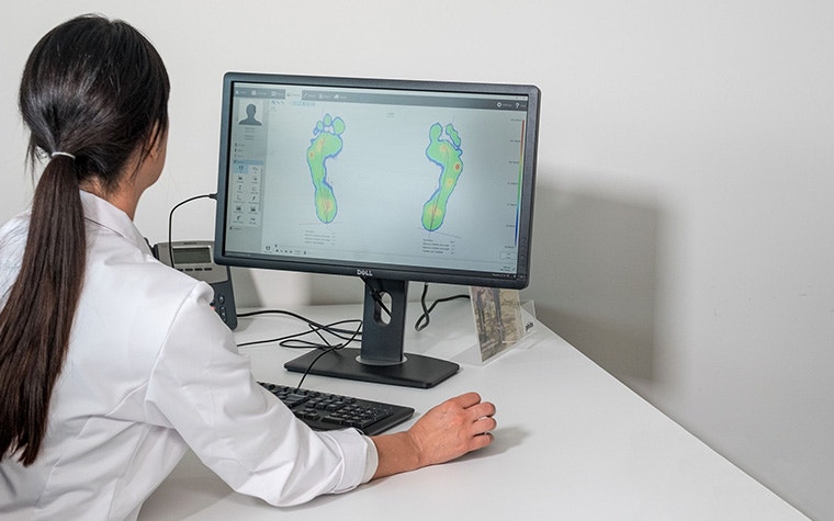 A foot health clinician in a white coat works at a computer showing the Footscan software, with pressure maps of a patient's feet.