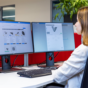 A woman looking at two computer screens showing 3D parts