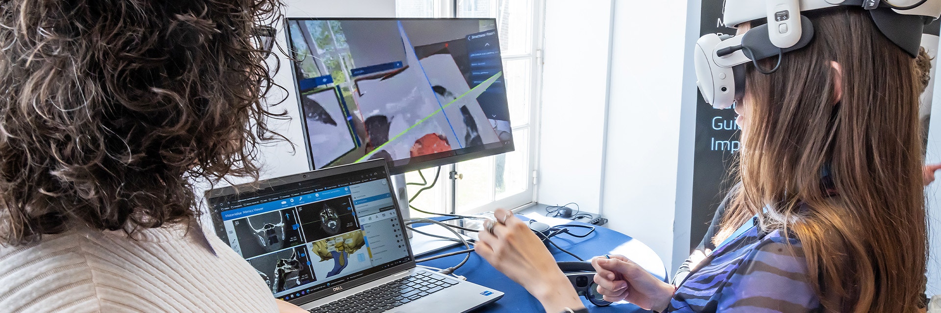 One woman wearing an XR headset at an event, holding out her hands to interact with a virtual model, while another watches on computer screens.