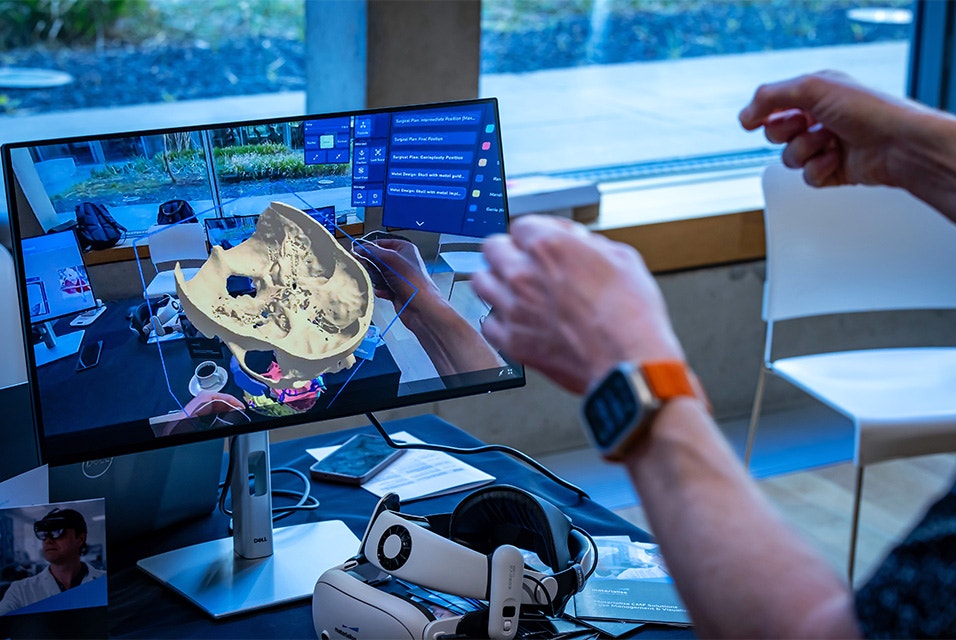 Person using hand gestures to interact with a 3D skull model via VR. A screen shows the skull model and a VR headset and controllers are on the desk.