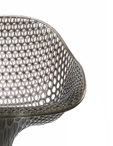 Close-up view of a geometrical, 3D-printed chair seat from a 3D-printed prototype of a customized, futuristic-looking wheelchair, using multiple 3D printing materials. The seat is lattice-structured and made of a translucent gray resin.