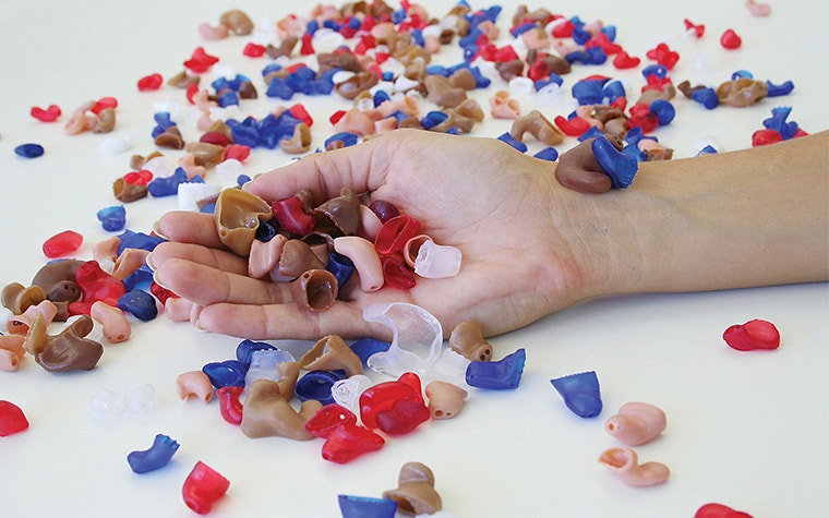 Hand holding 3D-printed hearing aids with many lying on the table around the hand