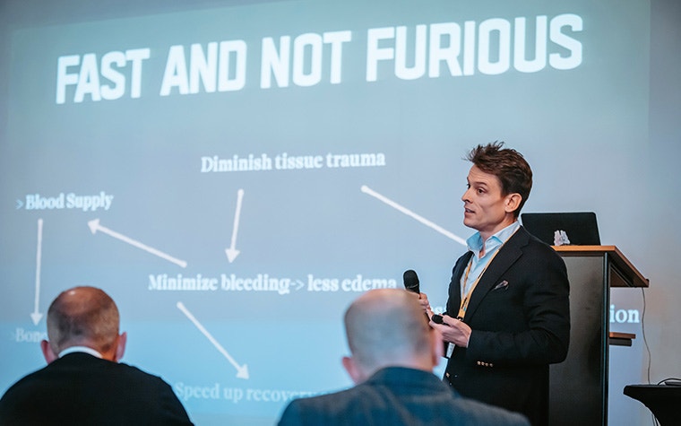 Dr. Javier Gutierrez speaking at the Materialise CMF Innovation Summit. The words "FAST AND NOT FURIOUS", "Diminish tissue trauma", "Blood supply", and "Minimize bleeding" show on the screen behind him, connected by arrows.
