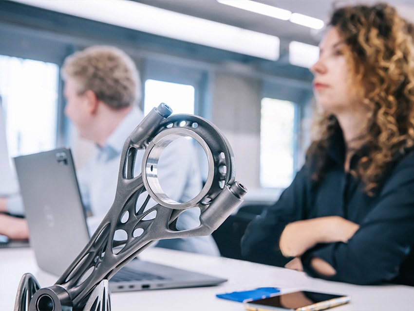 A metal 3D-printed part on a desk with two trainees sitting behind it on their laptops