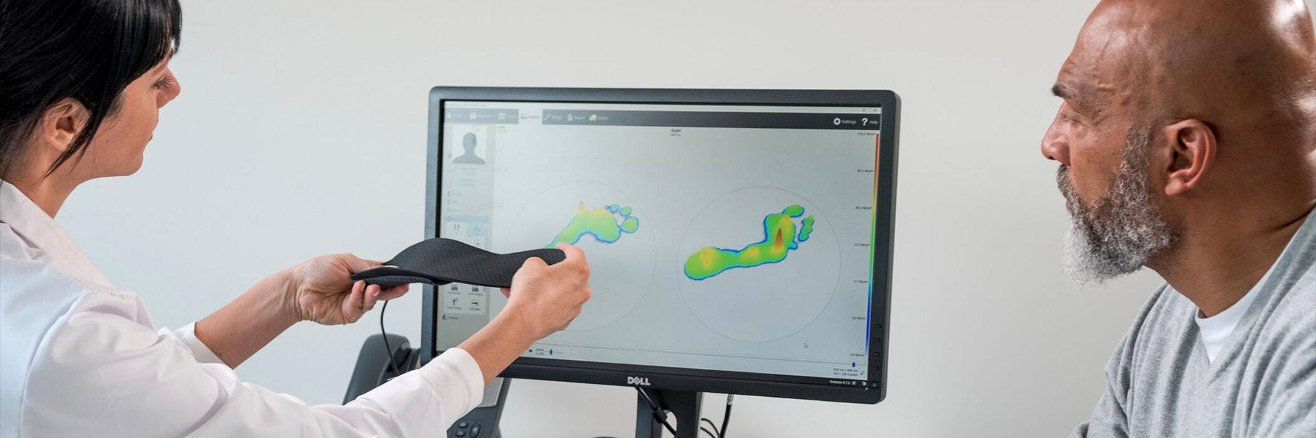 Doctor holding up a personalized insole while talking to a patient and looking at a screen with footscan software pulled up