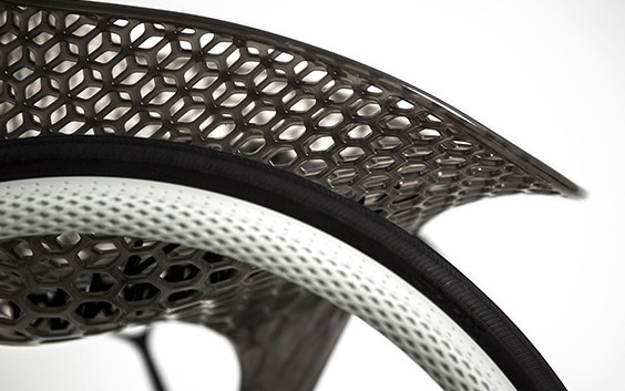 Close-up view of a geometrical, 3D-printed chair seat from a 3D-printed prototype of a customized, futuristic-looking wheelchair, using multiple 3D printing materials. The seat is lattice-structured and made of a translucent gray resin. The wheel spokes are made of 3D-printed metal.