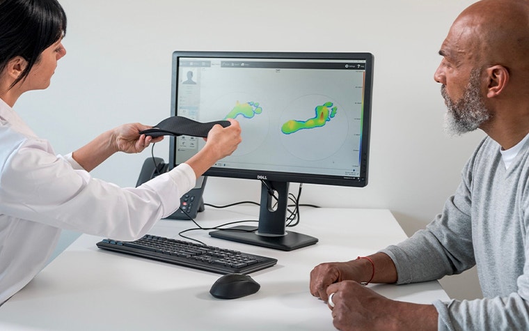 Doctor holding up a personalized insole while talking to a patient and looking at a screen with footscan software pulled up