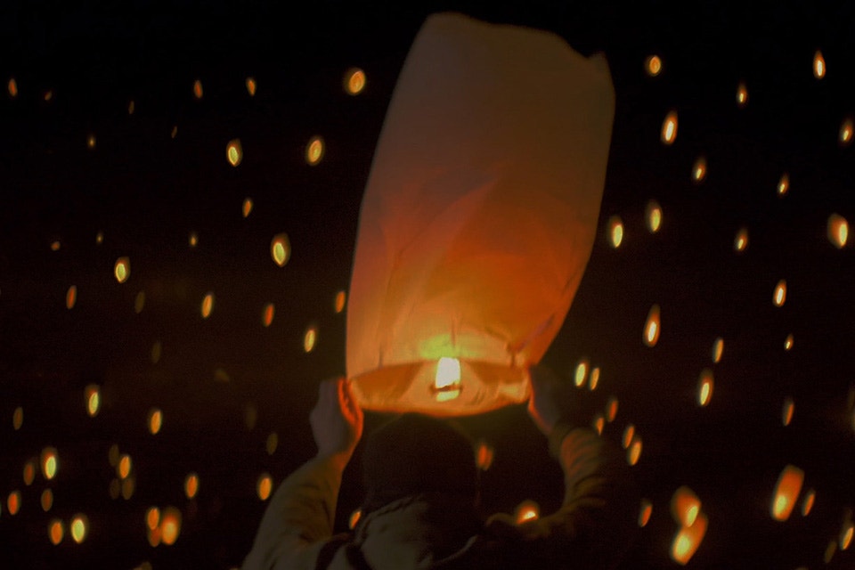 Person holding a lit paper lantern with many lanterns floating in the night sky in the background