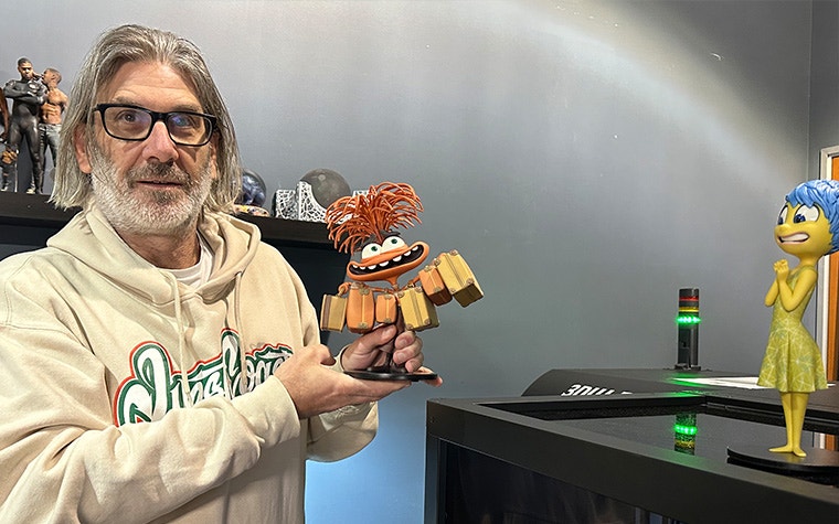 Jason Lopes from Gentle Giant Studios holding a 3D-printed model of Anxiety from Inside Out 2 with another model of Joy from the movie on a nearby 3D printer.