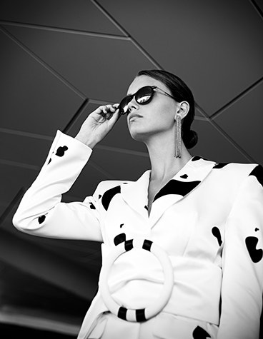Immagine in scala di grigi di una modella che guarda in alto mentre tiene in mano e indossa gli occhiali da sole della collezione Hoet Cabrio PR