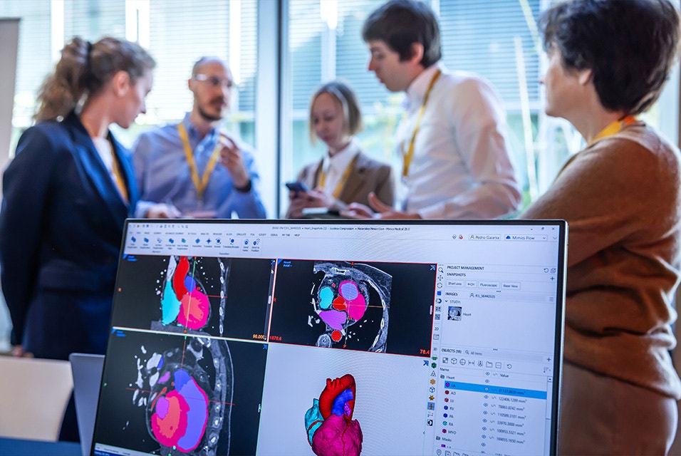 A computer screen displaying a segmented 3D heart model and medical imaging scans, with a group of 3D Planning and Printing in Hospitals Forum 2026 attendees talking in the background.