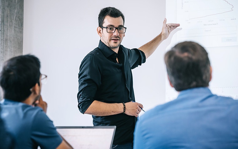 In a classroom, a man wearing a black shirt and glasses indicates a graph on a projector screen, watched on by two men in blue t-shirts.