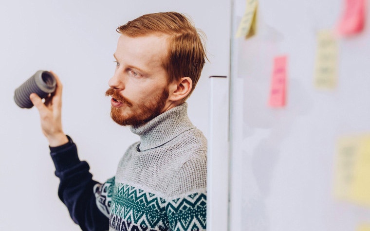 Man holding up a 3D-printed part and speaking next to a whiteboard with post-it notes attached