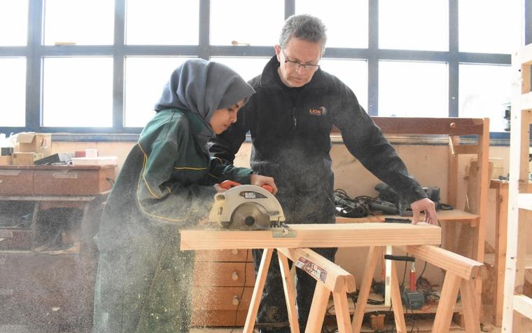 Woman using a saw on a piece of wood next to an instructor