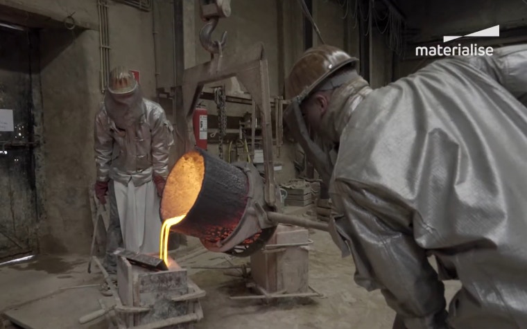 Two workers in a foundry are pouring molten bronze into a casting mold. Both workers are wearing full protective gear. 
