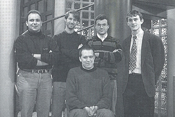 A black and white photo of four men standing and one man in a wheelchair
