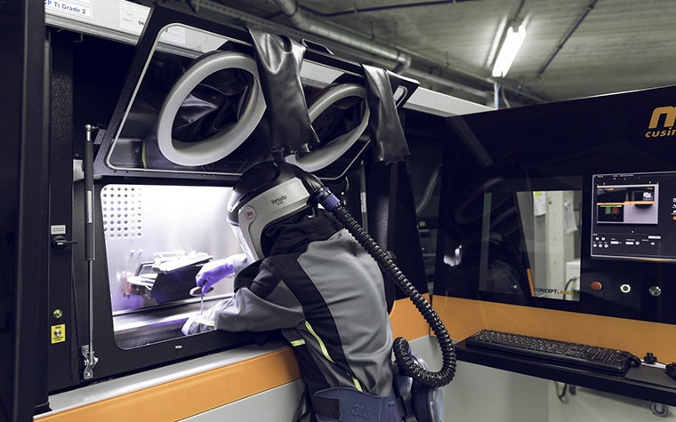 A man in protective gear removing a metal 3D-printed part from a printer.