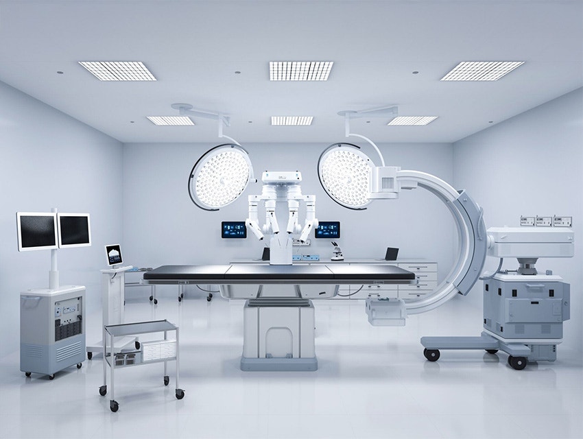A modern operating room with advanced medical equipment, including large overhead surgical lights, a robotic surgical system, dual monitors, and various mobile carts, all arranged around a central operating table in a sterile, brightly lit environment.