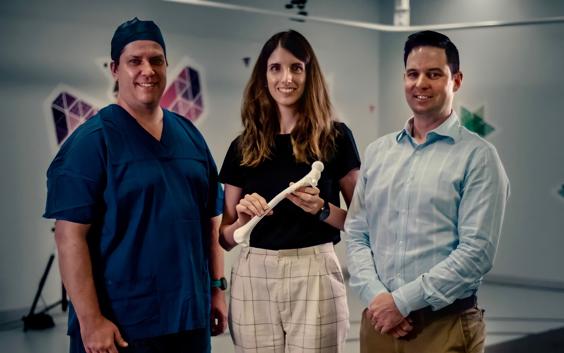 Three ADaPT team members smiling and posing with a 3D-printed bone model. One member is wearing surgical gear.