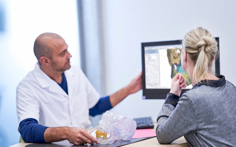 Surgeon showing a patient a digital model of their skull on a screen with a 3D-printed model of their skull on the table in between them