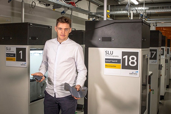 Process engineer Jonas Van Eyck stands in front of Materialise stereolithography machines holding 3D-printed parts 