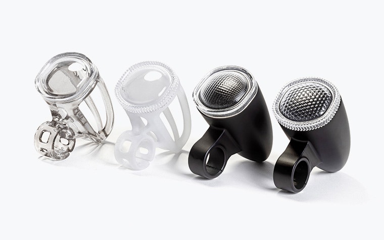 Four 3D-printed bicycle light housings made with VersaClear displayed in a row, showing different shades, from transparent and white prototypes to finished black units with textured reflective lenses.