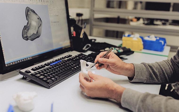 Hands checking the quality of a 3D-printed part at a desk in front of a screen showing the digital design of that part